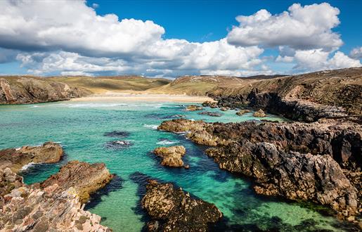 Rocky coastline with clear turquoise waters and sandy beach under a bright blue sky with scattered clouds. Rugged terrain surrounds the water.