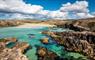 Rocky coastline with clear turquoise waters and sandy beach under a bright blue sky with scattered clouds. Rugged terrain surrounds the water.
